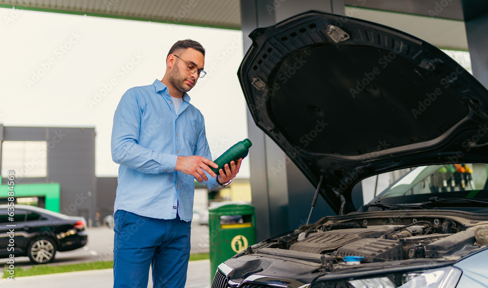 Fototapeta premium The man is standing in front of the raised hood, checking and changing the oil at the gas station
