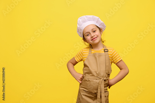 Fotografie Caucasian adorable little child girl in beige chef's apron and white hat, with hands on waist, smiles looking at camera, isolated yellow background