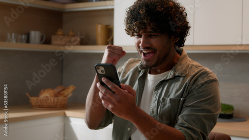Satisfied Indian male win with phone winning financial prize reward goal achievement with mobile app betting service online victory. Arabian happy man celebrating smartphone success at home kitchen photo