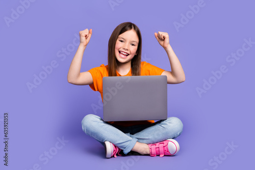 Full length photo of overjoyed schoolkid dressed orange t-shirt sitting with laptop raising fists isolated on purple color background © deagreez