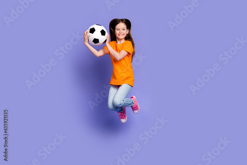 Full size photo of adorable active positive schoolgirl wear stylish t-shirt hold soccer ball jumping isolated on violet color background