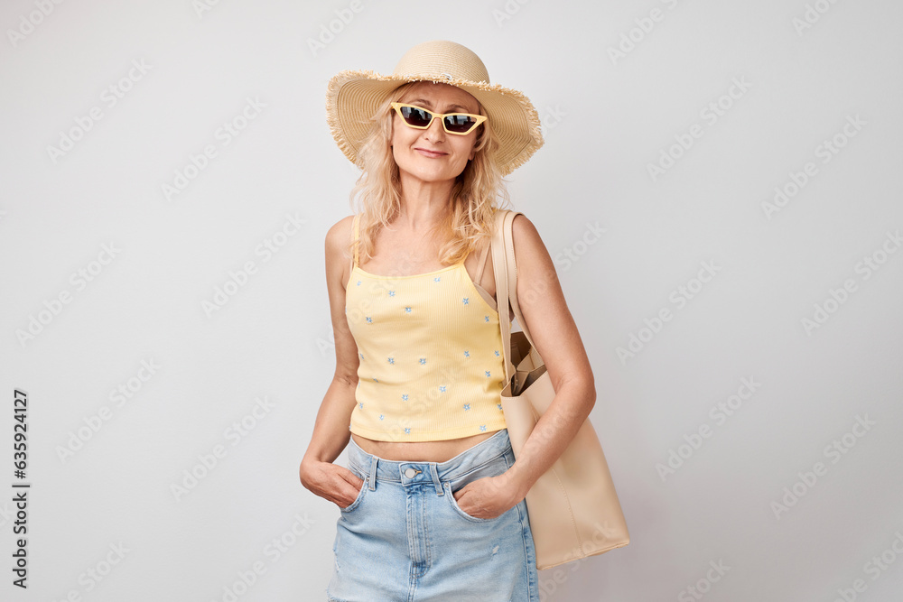 Portrait of happy beautiful blonde mature woman in yellow summer clothes, straw hat, sunglasses and shopping bag smiling isolated on white studio background. Vacation and travel concept Portrait of happy beautiful blonde mature woman in yellow summer clothes, straw hat, sunglasses and shopping bag smiling isolated on white studio background. Vacation and travel concept