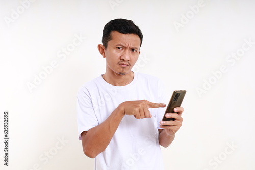 Curious asian funny man holding a cell phone while pointing on it. Isolated on white background