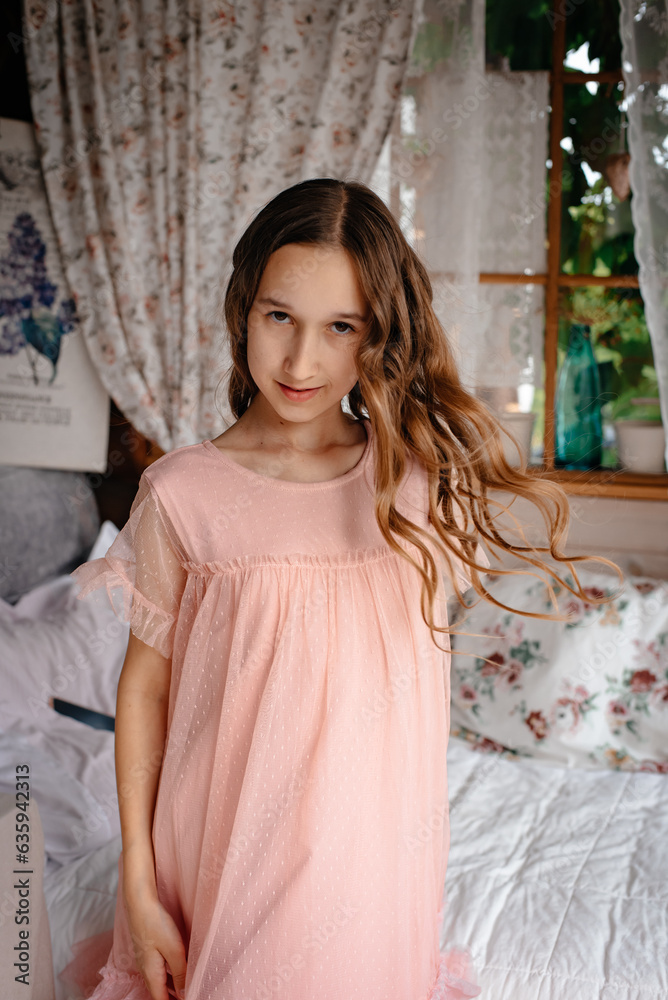 A little girl in a pink dress is sitting on a white bed and playing with her twisted brown hair