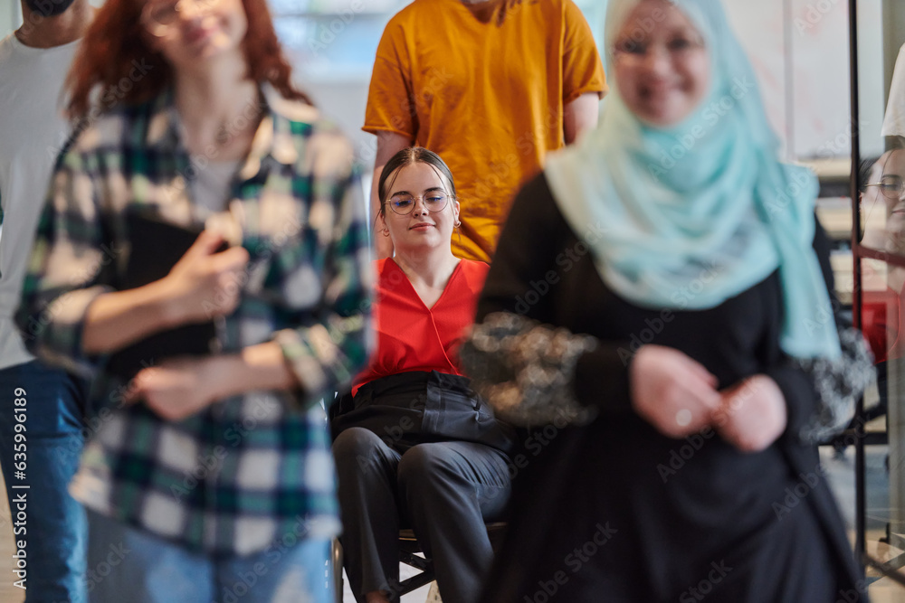 A diverse group of business people walking a corridor in the glass-enclosed office of a modern startup, including a person in a wheelchair and a woman wearing a hijab, showing a dynamic mix of