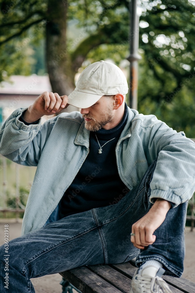 Fototapeta premium Young adult male is seated on a bench in a park, tying his baseball cap.