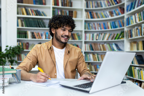Indian student with curly hair studying sitting among books on shelves, man watching video course writing in notebook smiling contentedly. photo