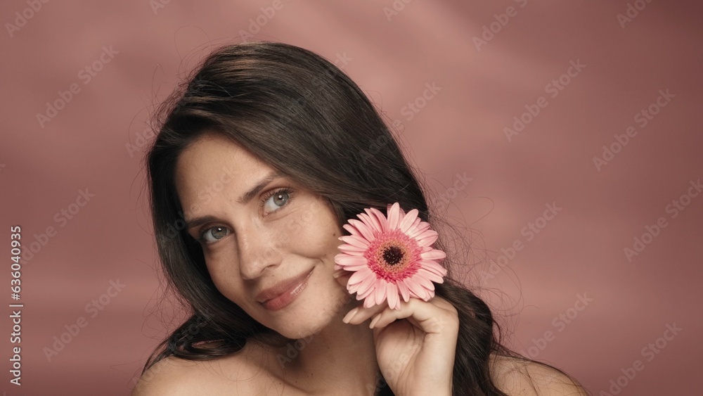 A woman touches a gerbera flower to her perfect skin of face. Portrait of a woman with a gerbera flower in the studio on a pink background close up. The concept of beauty, cosmetology, skin care. A woman touches a gerbera flower to her perfect skin of face. Portrait of a woman with a gerbera flower in the studio on a pink background close up. The concept of beauty, cosmetology, skin care.