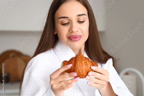 Beautiful young woman in bathrobe with tasty croissant at kitchen