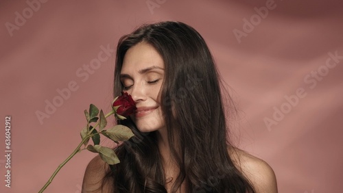 A seminude woman holds a red rose and inhales its fragrance. A woman in a studio on a pink background close up. Spring and summer inspiration. The concept of perfumery, cosmetics, skin care.