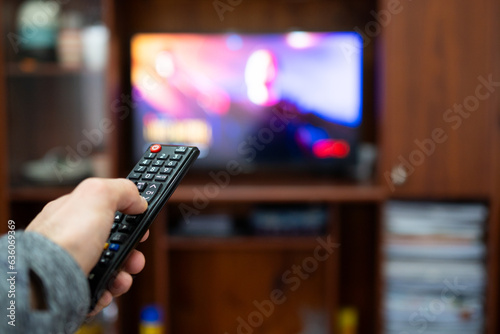 Hand of a person in the foreground holding a remote control and pointing to a TV in the background out of focus.