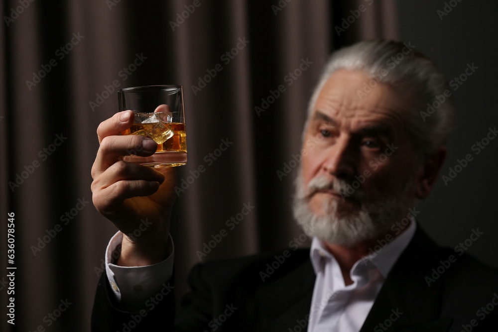 Senior man in suit holding glass of whiskey with ice cubes on brown background, selective focus