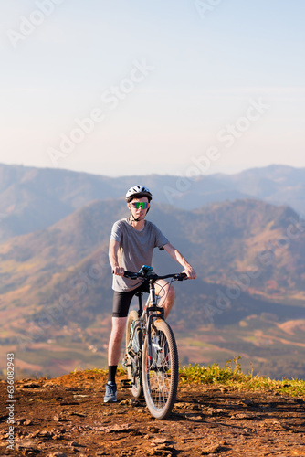 Cyclist resting atop his mountain bike, wearing sunglasses and a helmet, with a mountain landscape in the background