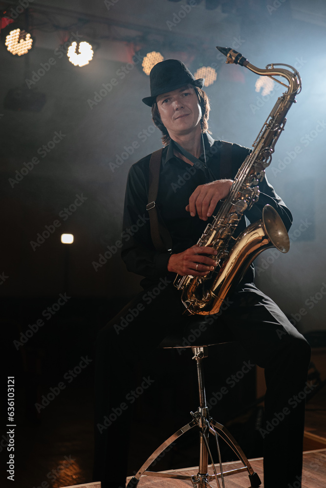 Naklejka premium Friendly man musician sitting on chair with saxophone in his hand and looking at camera