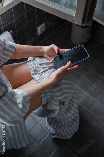 Faceless woman sitting on a toilet and scrolling her phone