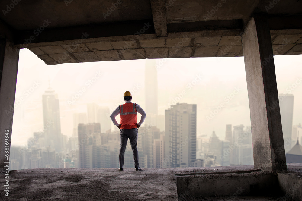 Back view of Indian male engineer wearing helmet and vest standing at construction site looking out for buildings and city view. Full length, Wide angle shot. Copy space. Real estate. Back view of Indian male engineer wearing helmet and vest standing at construction site looking out for buildings and city view. Full length, Wide angle shot. Copy space. Real estate.