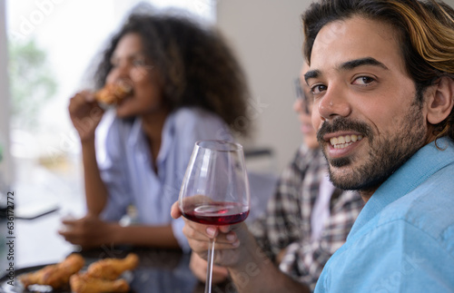 Portrait of smiling man holding glass of red wine while his friends sitting in background. Happy diversity people enjoying party at home 