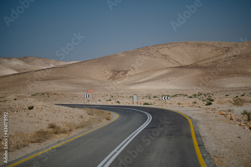 A bend in a lonely two-lane highway surrounded by a barren  hilly desert. Signage with arrows point to the left helps to guide drivers to stay on the road.