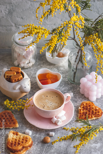 coffee with milk in a pink cup, cookies and a vase with yellow mimosa. Spring, stylish, delicate still life on a light background