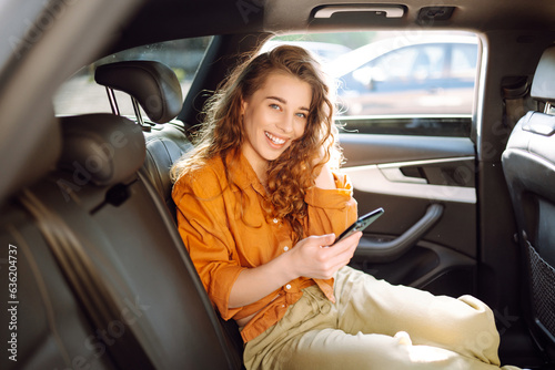 Cheerful woman with curly hair and stylish clothes sits in the back seat of a car, uses a mobile phone. The concept of business, technology, travel, online communication.