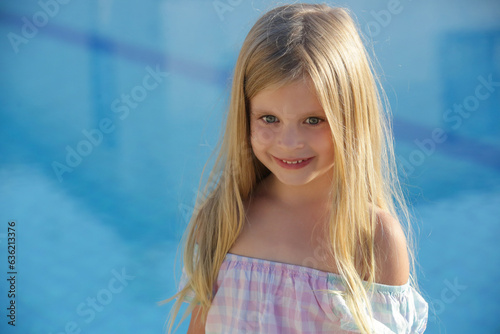 Portrait of adorable 5 years old girl with long blonde hair, blue eyes and blue swimming pool water behind 