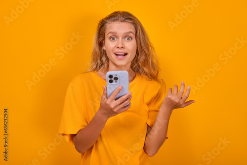 Young surprised optimistic Caucasian woman with phone in hand is experiencing positive shock after reading publication on social network or in personal blog of journalist stands on orange background.