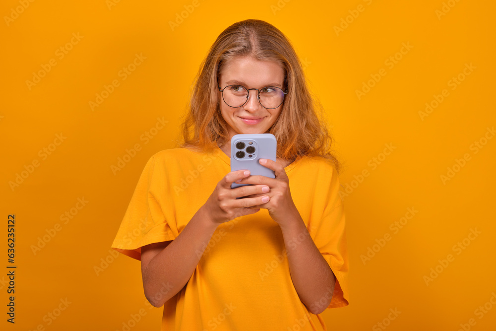Young cute shy Caucasian woman with phone in hands smiles sweetly and looks away getting to know guys on social networks or dating apps dressed in casual clothes stands in orange studio. Young cute shy Caucasian woman with phone in hands smiles sweetly and looks away getting to know guys on social networks or dating apps dressed in casual clothes stands in orange studio.