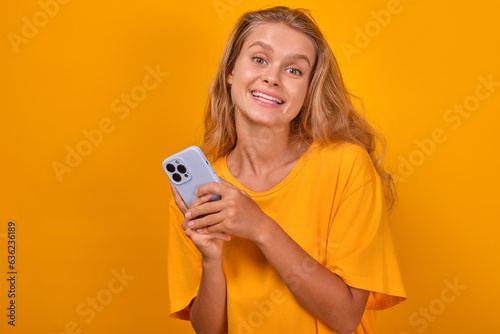Young satisfied casual Caucasian woman with mobile phone in hand looks at camera and uses smartphone to exchange messages with friends or communicate in instant messengers stands in orange studio.