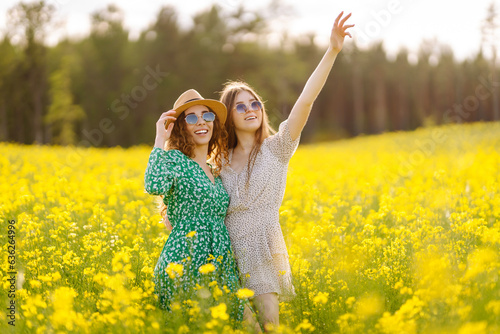 Two cheerful girlfriends dressed in stylish dresses are walking through a blooming rapeseed field. The concept of walking, active lifestyle, holidays.
