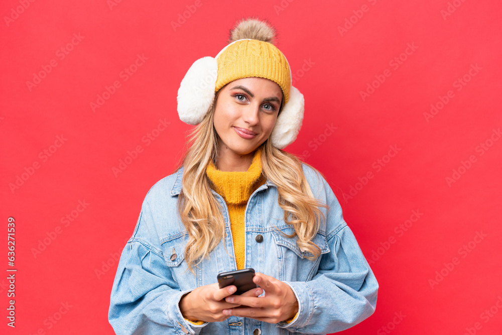Young pretty Uruguayan woman wearing winter muffs isolated on red background background sending a message with the mobile Young pretty Uruguayan woman wearing winter muffs isolated on red background background sending a message with the mobile