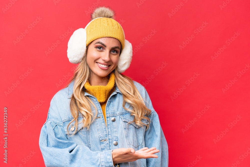 Young pretty Uruguayan woman wearing winter muffs isolated on red background background presenting an idea while looking smiling towards Young pretty Uruguayan woman wearing winter muffs isolated on red background background presenting an idea while looking smiling towards