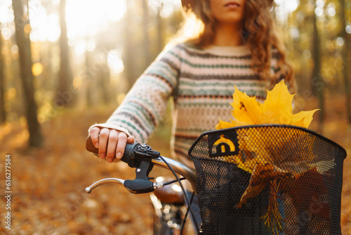 Fototapeta Naklejka Na Ścianę i Meble -  Smiling young woman in a hat and a stylish sweater with a bicycle walks and enjoys the autumn weather in the forest, among the yellow leaves at sunset.