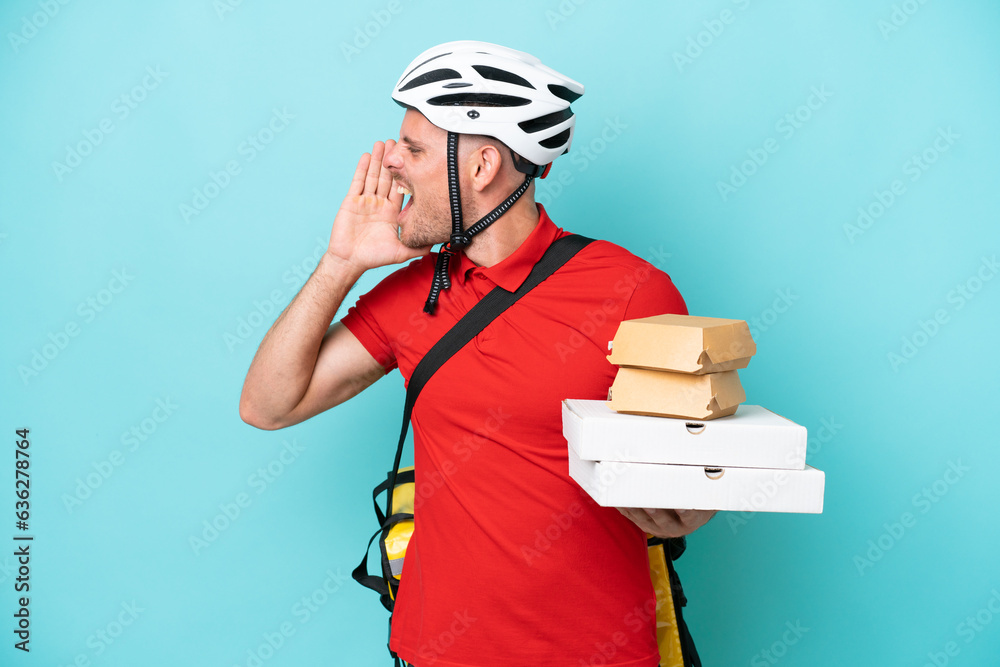 Young caucasian man with thermal backpack and holding fast food isolated on blue background shouting with mouth wide open to the side Young caucasian man with thermal backpack and holding fast food isolated on blue background shouting with mouth wide open to the side