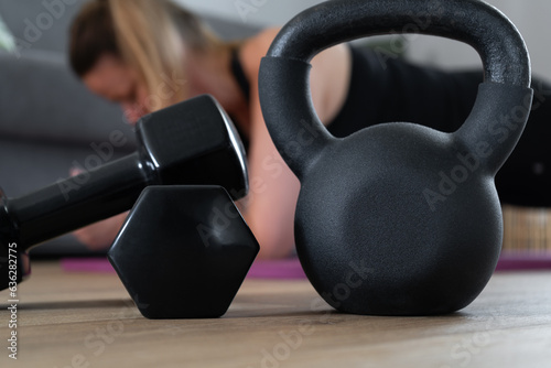 Woman exercising at home on yoga mat. Female doing plank exercise, working on abdominal muscles. House fitness workout, sport training concept. Focus on dumbbells and kettlebell gym equipment.