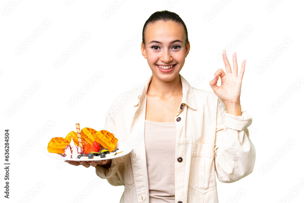 Young Arab woman holding waffles over isolated background showing ok sign with fingers Young Arab woman holding waffles over isolated background showing ok sign with fingers