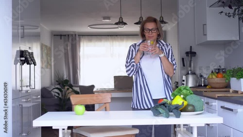 Healthy habit to drink water. Smiling middle age woman going to the table with glass of pure water with lemon standing on her kitchen and dancing at this monent. Control body hydration, Healthy living photo