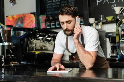 barista in apron talking on smartphone and using digital tablet on bar in coffee shop