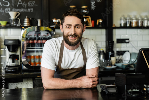 cheerful bearded barista looking at camera while standing near bar in coffee shop