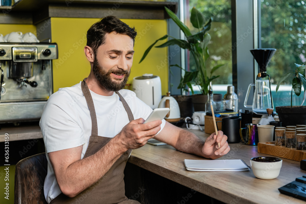 positive barista in apron using smartphone near notebook on worktop in coffee shop positive barista in apron using smartphone near notebook on worktop in coffee shop