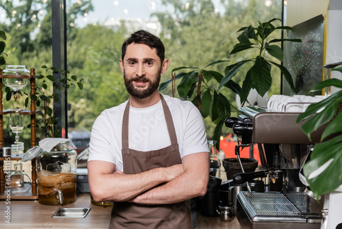 positive bearded barista in apron crossing arms and looking at camera while working in coffee shop