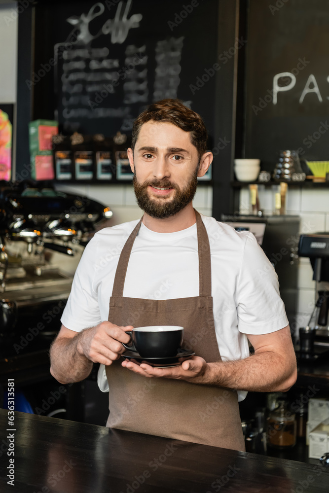 smiling bearded barista in apron holding cup of coffee and looking at camera in coffee shop smiling bearded barista in apron holding cup of coffee and looking at camera in coffee shop