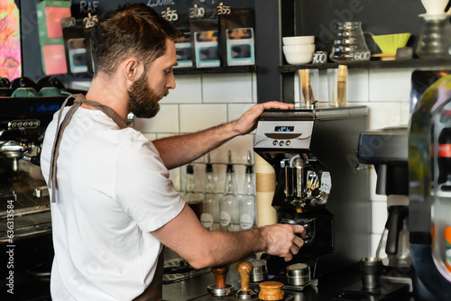 side view of barista in apron pouring coffee in holder near coffee machine while working in cafe