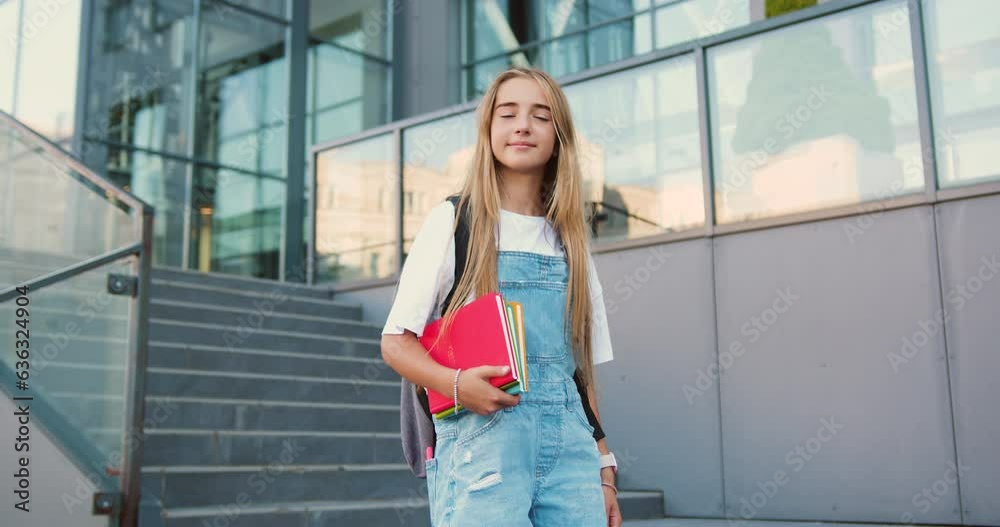 Portrait of lovely teen school girl with backpack smiling posing on camera near campus. Female elementary school student holds textbooks standing on stairs outside school building. Portrait of lovely teen school girl with backpack smiling posing on camera near campus. Female elementary school student holds textbooks standing on stairs outside school building.