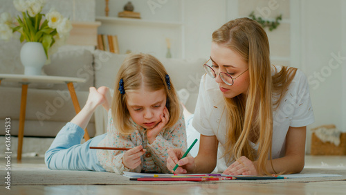 Caucasian mum nanny babysitter helping cute kid daughter teaching toddler child girl drawing picture with pencils lying on floor home learn creative art mother talking explain help draw happy family