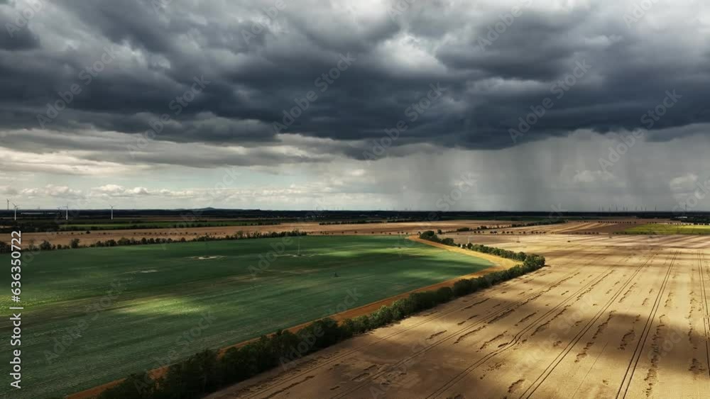 Dramatic dark sky and big rain clouds over the fields. Heavy rain in the distance. Wind turbines at the edge of the picture. Flight along cultivated fields. High voltage power line cuts through landsc