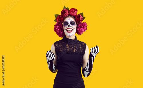 Positive emotions. Woman with sugar skull on Halloween rejoices in her success and victory, clenching her fists in joy. Woman with mexican face art and wreath on head, isolated on orange background.