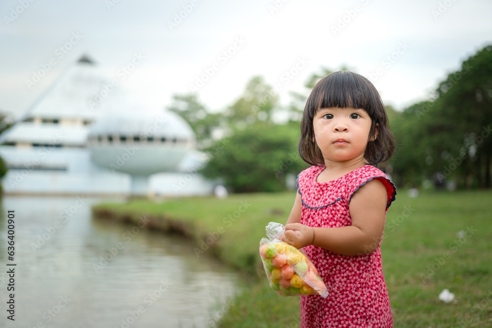 Naklejka premium A cute little kid in colorful pink dress is feeding bread to the fish in water pond at public park. Girl portrait photo with recreation activity. Selective focus.