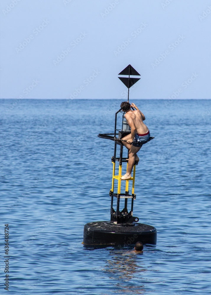 Obraz premium Teenage boys scaling a tall white buoy in the sparkling blue waters of Marsalforn, Gozo, Malta