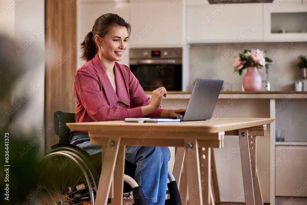 A smiling woman in a wheelchair working over a laptop at the home office. A smiling woman in a wheelchair working over a laptop at the home office.