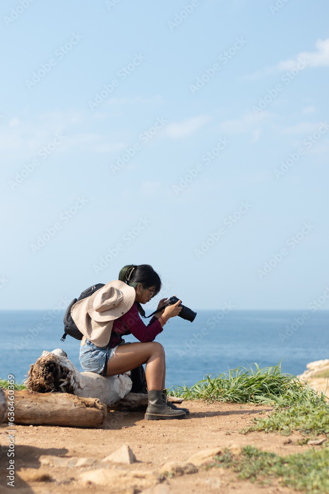 Young woman learning to use a camera by the sea while looking at the camera sitting on a log with the ocean in the background
